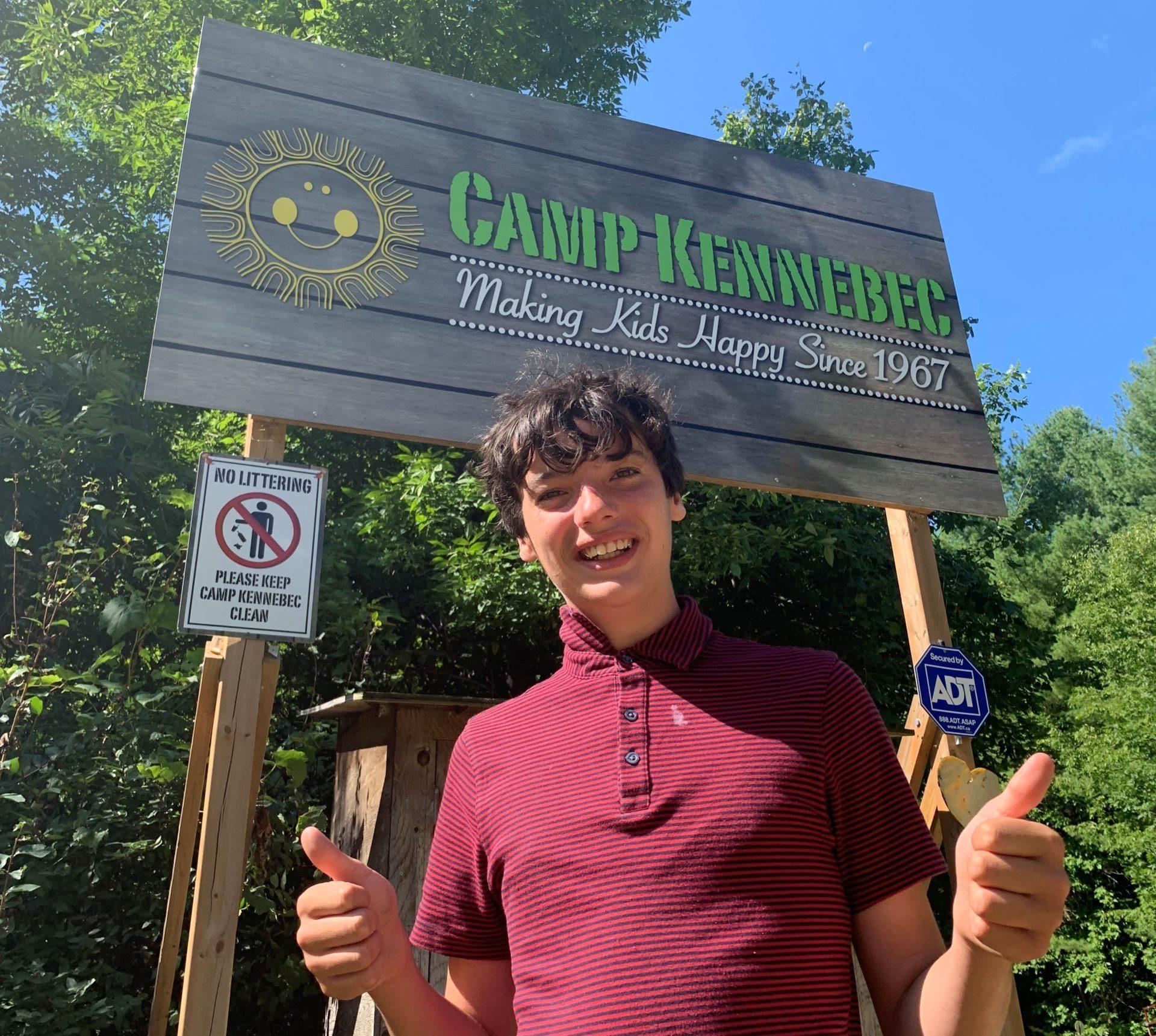 Male teen camper smiling and giving two thumbs up in front of the Camp Kennebec sign.