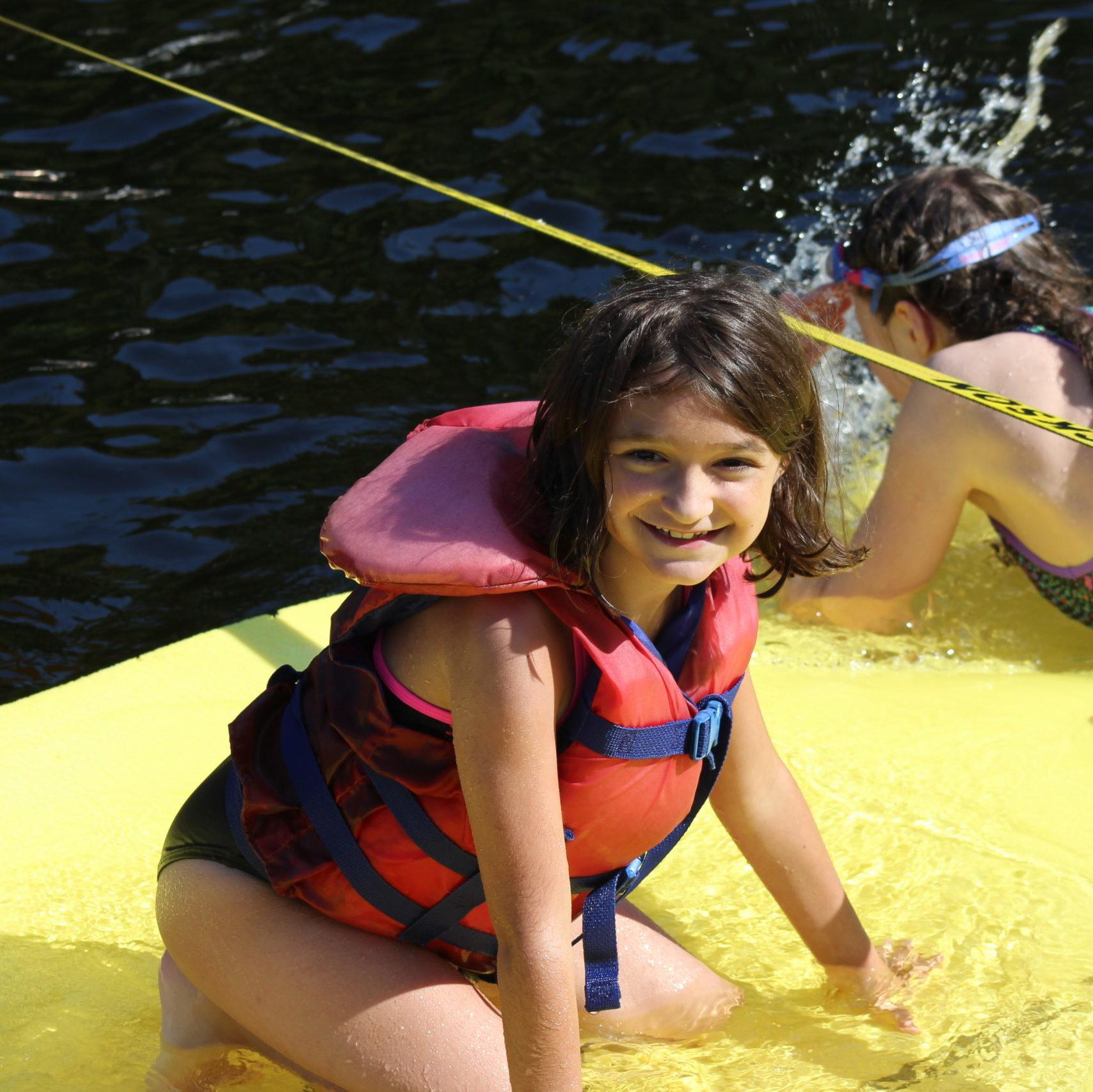 Young female camper wearing a life jacket smiling on the yellow floating mat at Camp Kennebec