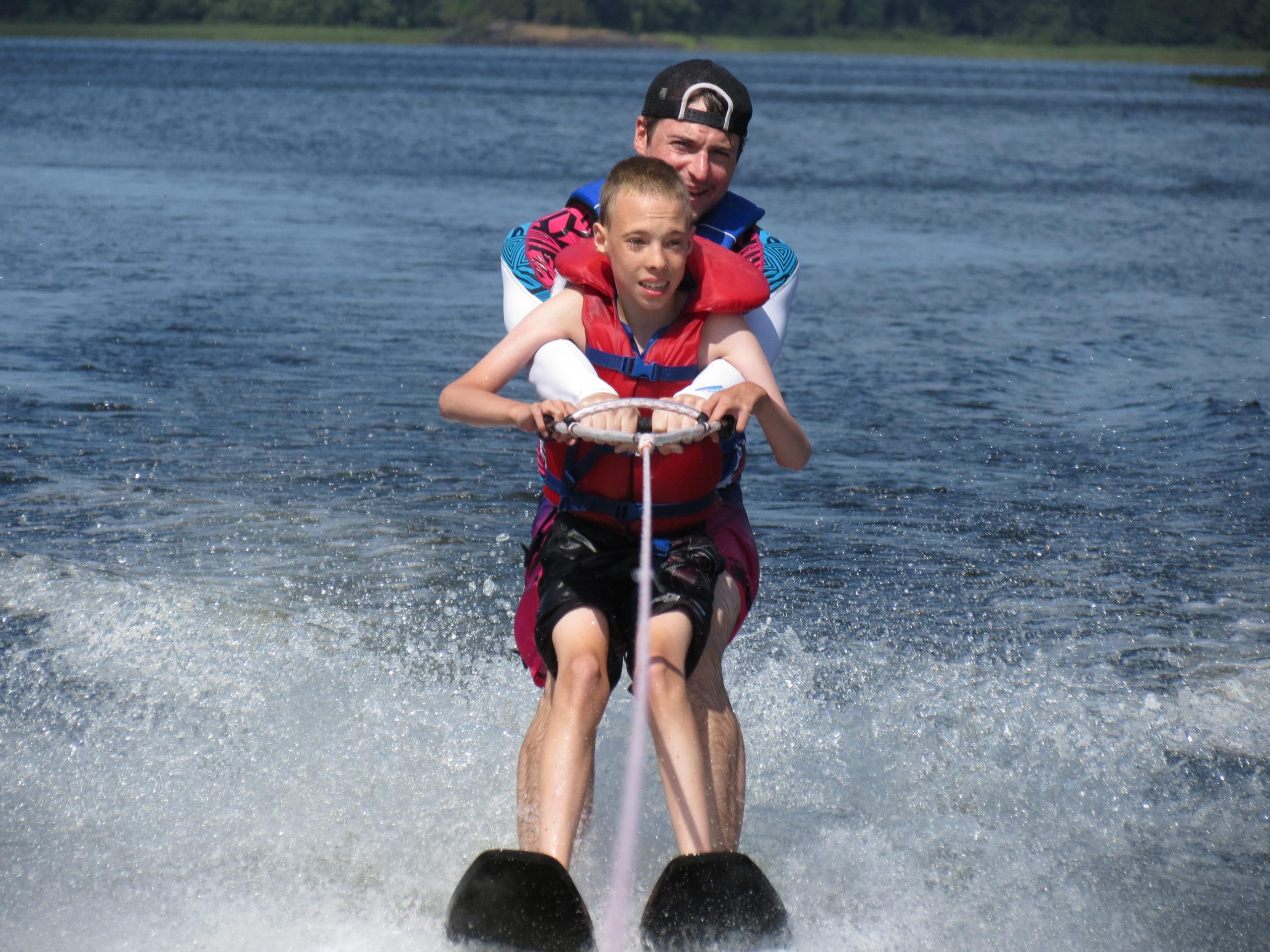 Young male camper learning to waterski with counsellor behind him holding him up at Camp Kennebec